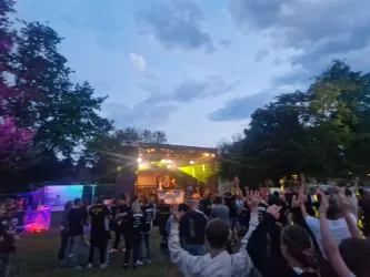 Ein lebhaftes Musikfestival im Freien mit einer großen Menschenmenge. Die Bühne ist hell beleuchtet und von Bäumen umgeben.