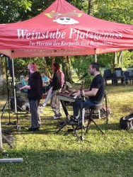 Eine Musikgruppe spielt unter einem roten Zelt im Freien. Im Hintergrund sind Stühle und Bäume sichtbar.