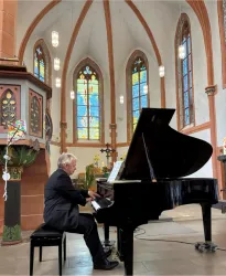 Ein Mann spielt Klavier in einer Kirche mit bunten Fenstern. Der Raum hat eine ruhige und feierliche Atmosphäre.