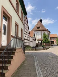 Eine malerische Straße mit Fachwerkhäusern und einem kleinen Platz. Im Hintergrund ist eine charmante Kapelle zu sehen.
