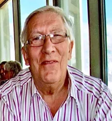 Ein älterer Mann mit Brille und gestreiftem Hemd lächelt freundlich. Im Hintergrund sind verschwommene Gestalten und große Fenster zu sehen.