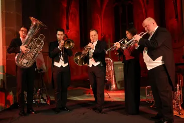 Eine Band spielt Live-Musik auf einer Bühne. Fünf Musiker mit verschiedenen Blasinstrumenten sind in eleganter Kleidung abgebildet.