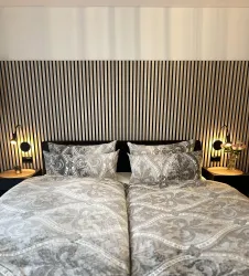 Ein modernes Schlafzimmer mit einem Doppelbett und elegantem, gemustertem Bettzeug. Die Wände sind mit schmalen, gestreiften Tapeten dekoriert und beleuchtet von stilvollen Nachttischlampen.