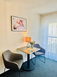 Ein gemütlicher Raum mit einem Tisch und zwei Stühlen. Auf dem Tisch steht eine Lampe und ein Blumenstrauß, im Hintergrund hängt ein farbenfrohes Bild.