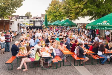 Eine belebte Veranstaltung mit vielen Menschen, die an langen Tischen sitzen. Überdachte Sitzbereiche und Bäume schaffen eine einladende Atmosphäre.