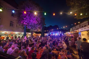 Ein belebtes Fest mit vielen Menschen unter bunten Lichtern. Stände und eine fröhliche Atmosphäre prägen die Nacht.