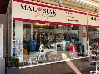 Ein elegantes Schaufenster eines Juweliergeschäfts mit verschiedenen Taschen, Schmuck und Uhren. Die Beschriftung betont die angebotenen Produkte wie Uhren und Geschenkartikel.