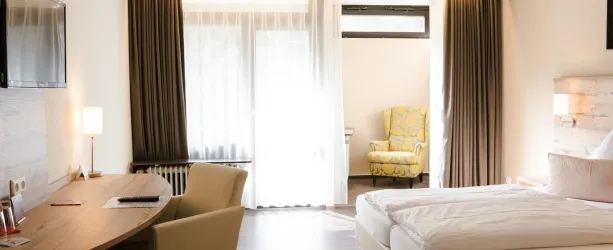 Ein modernes Hotelzimmer mit einem Einzelbett, einem Schreibtisch und einem gemütlichen Sessel. Große Fenster sorgen für viel Tageslicht.