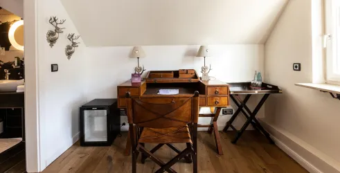 Ein gemütliches Arbeitszimmer mit einem alten Schreibtisch und einer Holzstuhl. An der Wand sind dekorative Elemente und eine große Fensterquelle für natürliches Licht.