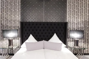 Ein elegantes Schlafzimmer mit einem schwarzen gepolsterten Kopfteil und stilvollen Lampen. Die Wände sind mit einem modernen Muster dekoriert.