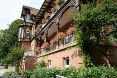 Ein elegantes Haus mit Balkonen, die mit bunten Blumen geschmückt sind. Es ist von einem grünen Garten umgeben und zeigt eine charmante Architektur.