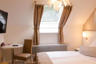 Ein gemütliches Zimmer mit einem Bett, einem Tisch und einem Stuhl. Große Fenster mit Vorhängen lassen viel Licht herein.