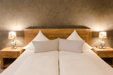 Ein gemütliches Doppelbett mit weiße Bettwäsche und zwei Kissen. Neben dem Bett stehen Nachtkommoden mit dekorativen Lampen.