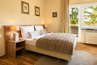 Ein gemütliches Schlafzimmer mit einem großen Bett und warmen Holzfarben. Es gibt große Fenster, die viel Tageslicht hereinlassen.