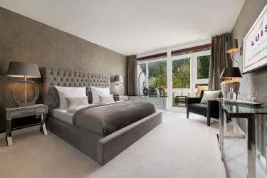 Ein modernes Schlafzimmer mit einem großen, gepolsterten Bett und stilvollen Möbeln. Große Fenster bieten einen Blick auf die grüne Umgebung.