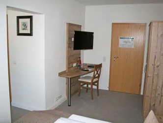 Ein einfaches Zimmer mit einem Tisch, einem Stuhl und einem Fernseher. Die Wände sind hell gestrichen und es gibt eine Holzverkleidung.