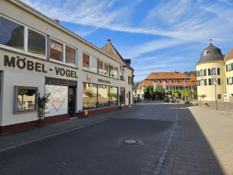 Eine ruhige Straße mit einem Möbelgeschäft namens "Möbel-Vogel". Im Hintergrund sind charmante Gebäude und ein blauer Himmel zu sehen.