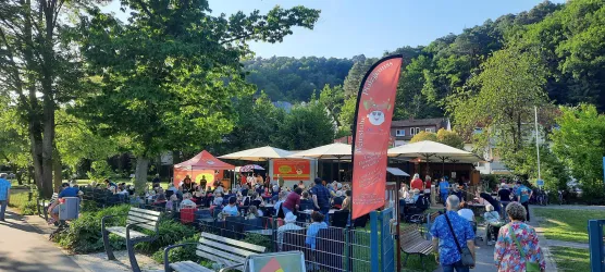 Eine belebte Außenterrasse mit vielen Gästen unter Sonnenschirmen. Umgeben von Bäumen und einem grünen Park.
