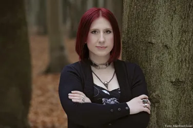 Eine Person mit rotem Haar steht an einem Baum im Wald. Der Boden ist mit braunen Blättern bedeckt.