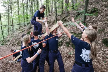 Eine Gruppe von jungen Menschen arbeitet zusammen, um eine Aufgabe im Wald zu lösen. Sie helfen sich gegenseitig, während sie an einem Seil ziehen und eine Person auf ihren Schultern balanciert.