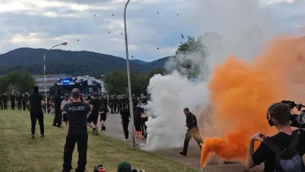 Mehrere Polizisten stehen in einer Konfrontationssituation. Orangefarbener Rauch und Explosionen sind sichtbar, während im Hintergrund eine Polizeifahrzeug und eine Gruppe von Einsatzkräften zu sehen sind.