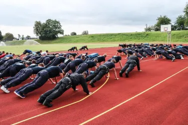 Eine Gruppe von Menschen macht Liegestütze auf einem Sportplatz. Im Hintergrund sind Bäume und ein Basketballkorb sichtbar.
