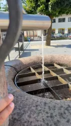 Ein Wasserbrunnen mit einem Edelstahl-Auslauf, aus dem Wasser in einen runden Behälter fließt. Im Hintergrund sind Bäume und Gebäude sichtbar.