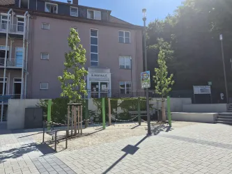 Ein modernes Gebäude mit einer Eingangszone und Grünflächen. Es gibt einige Bäume und Sitzgelegenheiten im Freien.