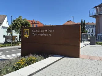 Eine moderne Hinweistafel mit dem Namen "Martin-Butzer-Platz" in einer städtischen Umgebung. Im Hintergrund sind Wohnhäuser und ein blauer Himmel zu sehen.