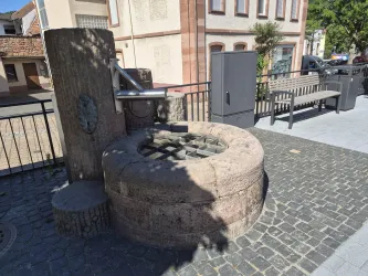 Ein historischer Brunnen aus Stein steht auf einem gepflasterten Platz. Im Hintergrund sind einige Gebäude und eine Bank zu sehen.