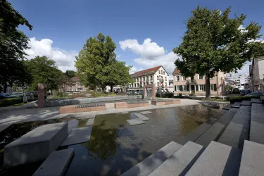 Ein attraktiver Platz mit Wasserbecken und modernen Steinbänken. Im Hintergrund sind Bäume und Gebäude zu sehen.
