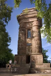 Ein großer, historischer Turm aus Stein, umgeben von Bäumen. Zwei Personen mit Fahrrädern stehen davor und genießen die Umgebung.