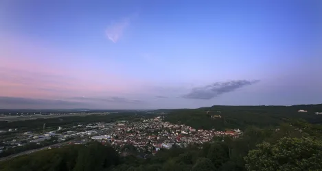 Ein schöner Ausblick auf eine Stadt in der Abenddämmerung. Der Himmel ist in sanften Farben gefärbt und die Umgebung ist grün und bewaldet.