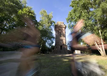 Ein historischer Turm umgeben von Bäumen. Im Vordergrund sind verschwommene Radfahrer zu sehen, die vorbeifahren.