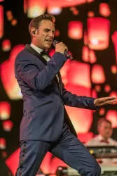 Ein Sänger in einem eleganten Anzug steht auf der Bühne und hält ein Mikrofon. Im Hintergrund leuchten farbige Lichter und schaffen eine lebhafte Atmosphäre.