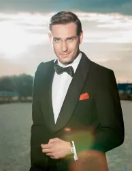 Ein eleganter Mann in einem Smoking mit einer Fliege steht im Freien. Er lächelt und der Sonnenuntergang im Hintergrund sorgt für eine romantische Atmosphäre.