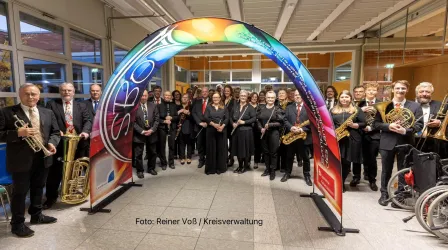 Eine große Gruppe von Musikern steht unter einem bunten Bogen. Sie tragen schwarze Kleidung und halten verschiedene Instrumente in den Händen.