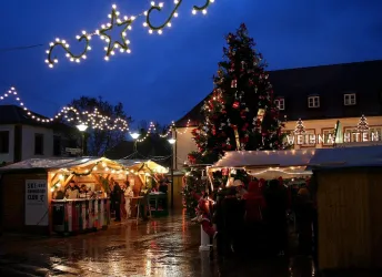 Ein Weihnachtsmarkt bei Abenddämmerung mit festlich beleuchteten Ständen und einem großen geschmückten Baum. Die Atmosphäre ist gemütlich und einladend.