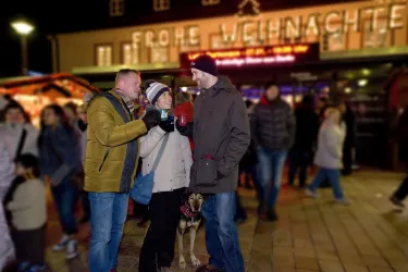 Drei Personen stehen auf einem Weihnachtsmarkt und halten Getränke in den Händen. Im Hintergrund ist ein beleuchtetes Schild mit der Aufschrift „Frohe Weihnachten“ zu sehen.