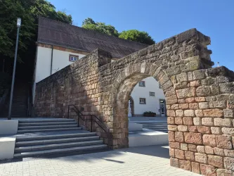Eine historische Steinmauer mit einem Torbogen, umgeben von modernem Treppenbereich. Im Hintergrund ist ein Gebäude mit weißen Wänden und einem begrünten Hang zu sehen.