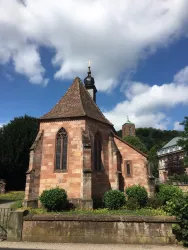 Eine kleine Kirche aus rotem Sandstein mit einem spitzen Dach und einem erhöhten Turm. Der Himmel ist klar mit einigen Wolken, umgeben von grünen Sträuchern und Bäumen.