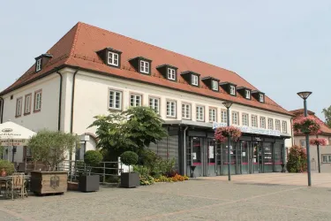 Ein modernes Gebäude mit einem roten Satteldach und großen Fenstern. Vor dem Haus gibt es Pflanzen und Blumen, die den Platz verschönern.
