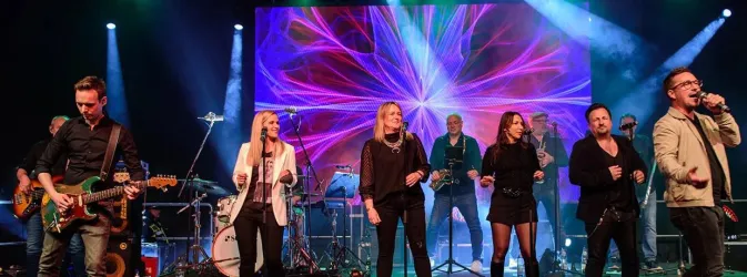 Eine Liveband steht auf der Bühne und spielt Musik. Bunte Lichter und eine dynamische Hintergrundprojektion schaffen eine fröhliche Atmosphäre.