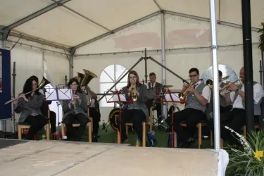 Eine Blaskapelle spielt in einem Zelt. Die Musikerinnen und Musiker sind in formellen Anzügen gekleidet und stehen auf der Bühne.