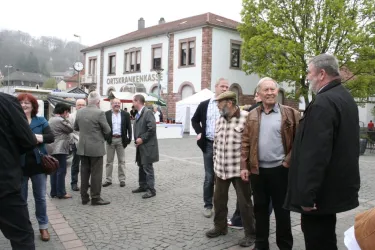 Eine Gruppe von Menschen versammelt sich auf einem Platz. Im Hintergrund ist ein Gebäude mit der Aufschrift "ORTSkrankenkasse" zu sehen.