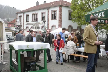 Ein belebter Marktplatz mit Menschen, die miteinander sprechen und sich unterhalten. Im Hintergrund sind Stände und ein Gebäude mit dem Schriftzug "ORTSKRANKENKASS" zu sehen.