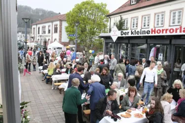 Eine belebte Straße mit vielen Menschen, die an Tischen sitzen und essen. Einige Gäste feiern mit Luftballons und eine freundliche Atmosphäre ist zu spüren.