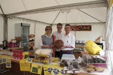 Ein Markstand mit drei Personen, die verschiedene Backwaren präsentieren. Im Hintergrund sind weitere Produkte und ein Banner mit der Aufschrift "Napoli" zu sehen.