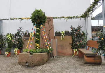 Ein dekorierter Brunnen mit bunten Ostereiern und grünen Pflanzen. Im Hintergrund sind weitere Pflanzen und eine Bank zu sehen.