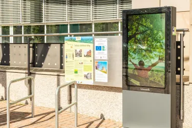 Ein Werbeschild mit einem Bildschirm und einem plakatartigen Infostand. Im Hintergrund sind Fenster und eine hell beleuchtete Wand zu sehen.